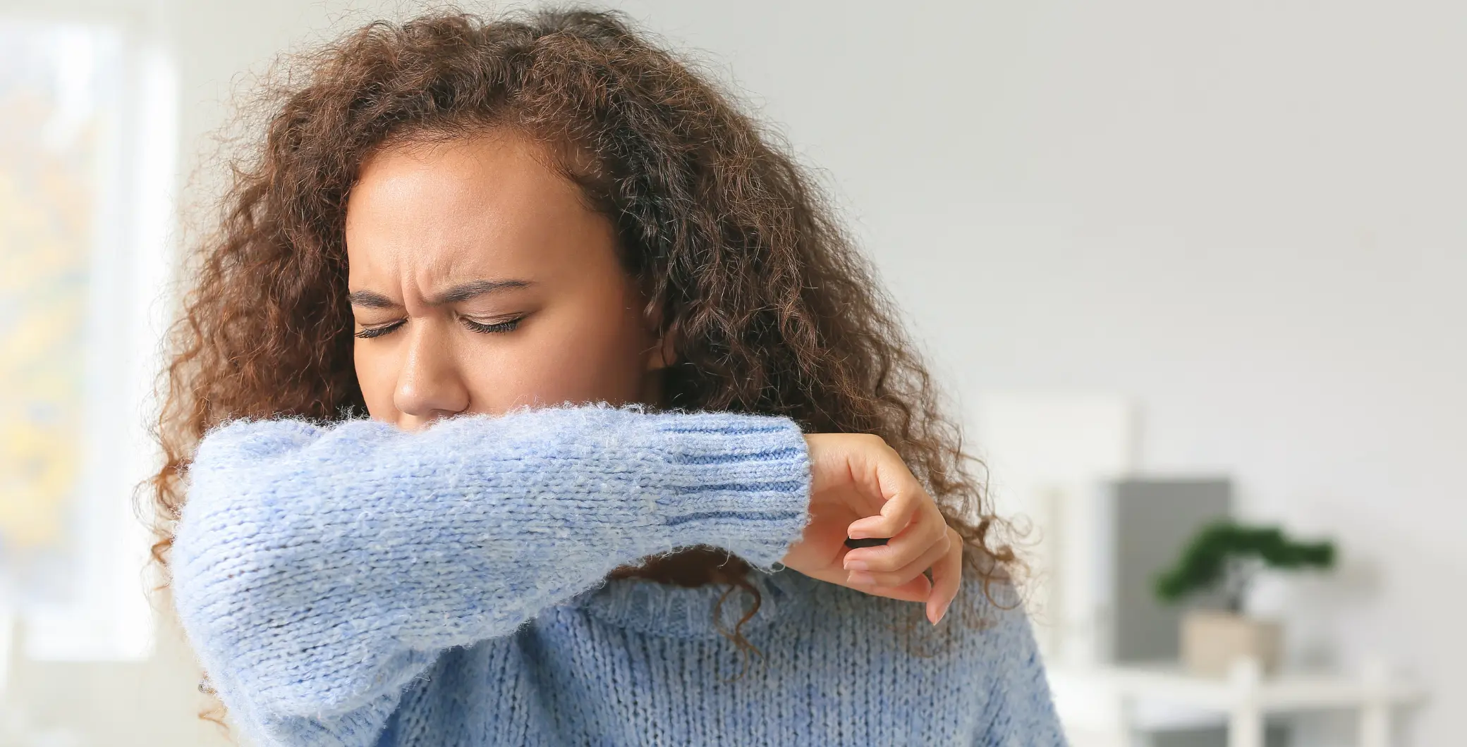 Young woman with curly hair sneezing into her elbow, wearing a blue sweater, indoors with a blurred background.