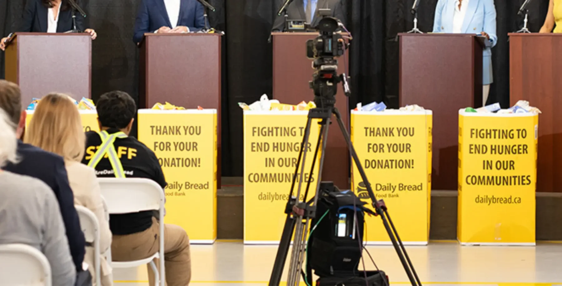 People attending a charity event with donation boxes and a camera setup, promoting hunger relief in communities, at an indoor venue.