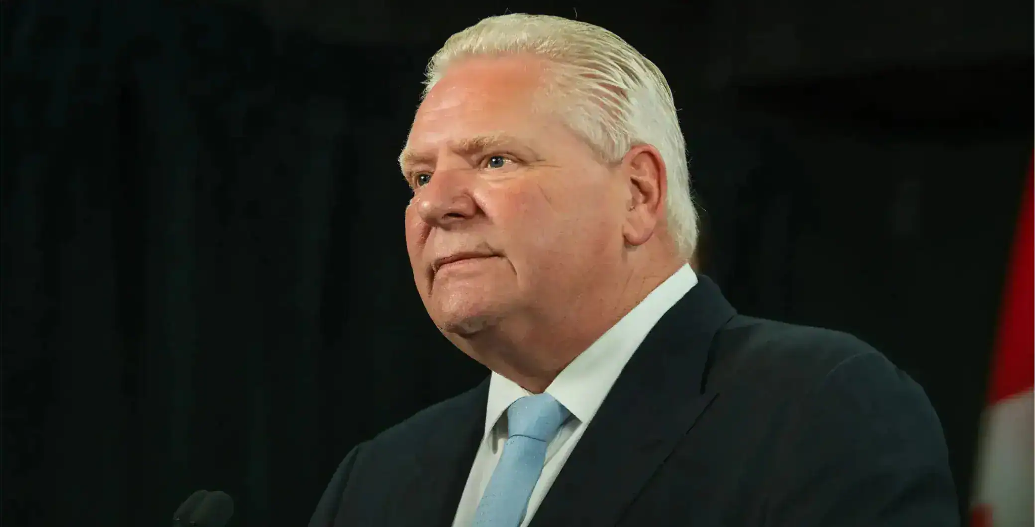 A serious-looking man with white hair in a dark suit and light blue tie, standing in front of a black background, possibly at a formal event or press conference.