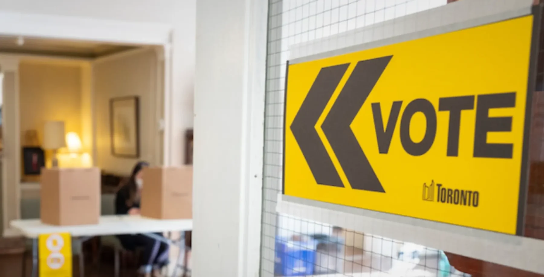 Toronto voting station with a large yellow and black Vote sign near a window, with a person and voting booths visible inside.
