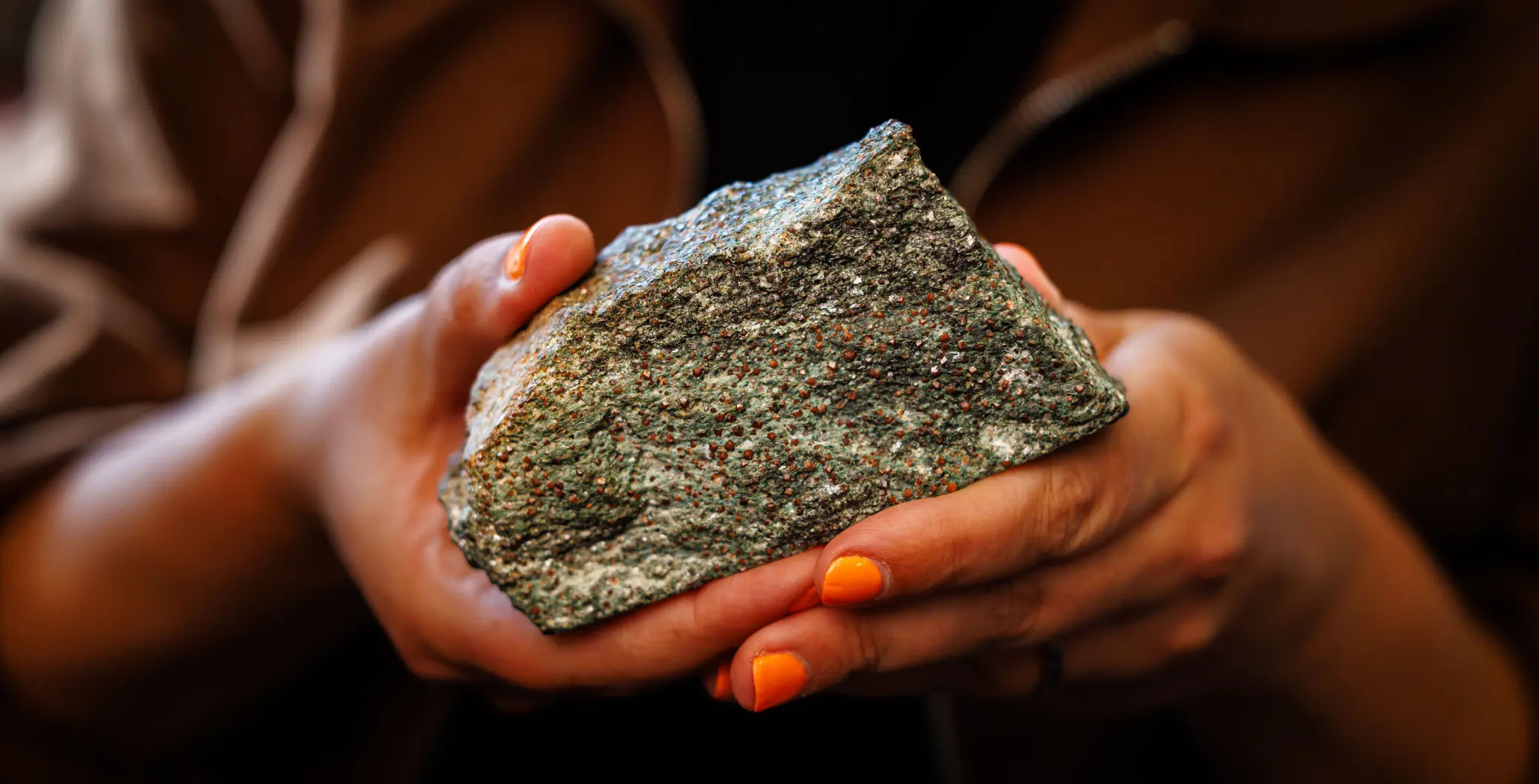 Close-up of a person holding a rough, greenish rock with both hands, with a blurred natural background.