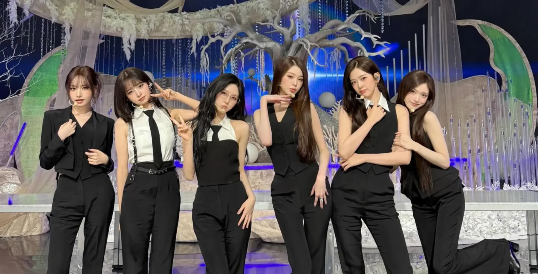 Six women in black outfits perform on stage with a fantasy-themed backdrop featuring a large tree and moon.