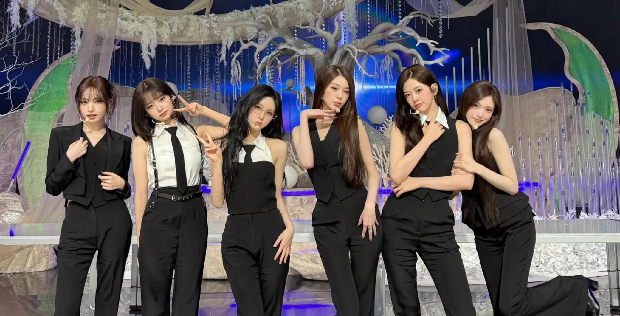Six women in black outfits perform on stage with a fantasy-themed backdrop featuring a large tree and moon.