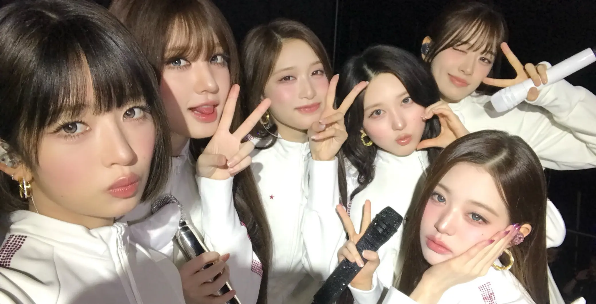 Group of six young women posing together, making peace signs and smiling, in a casual indoor setting.