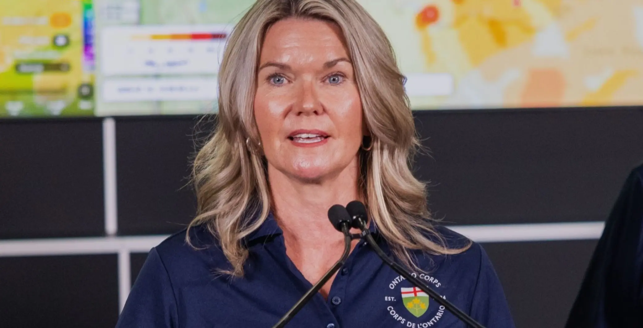 Ontario government official speaking at a press conference with a blurred background of screens and a microphone.