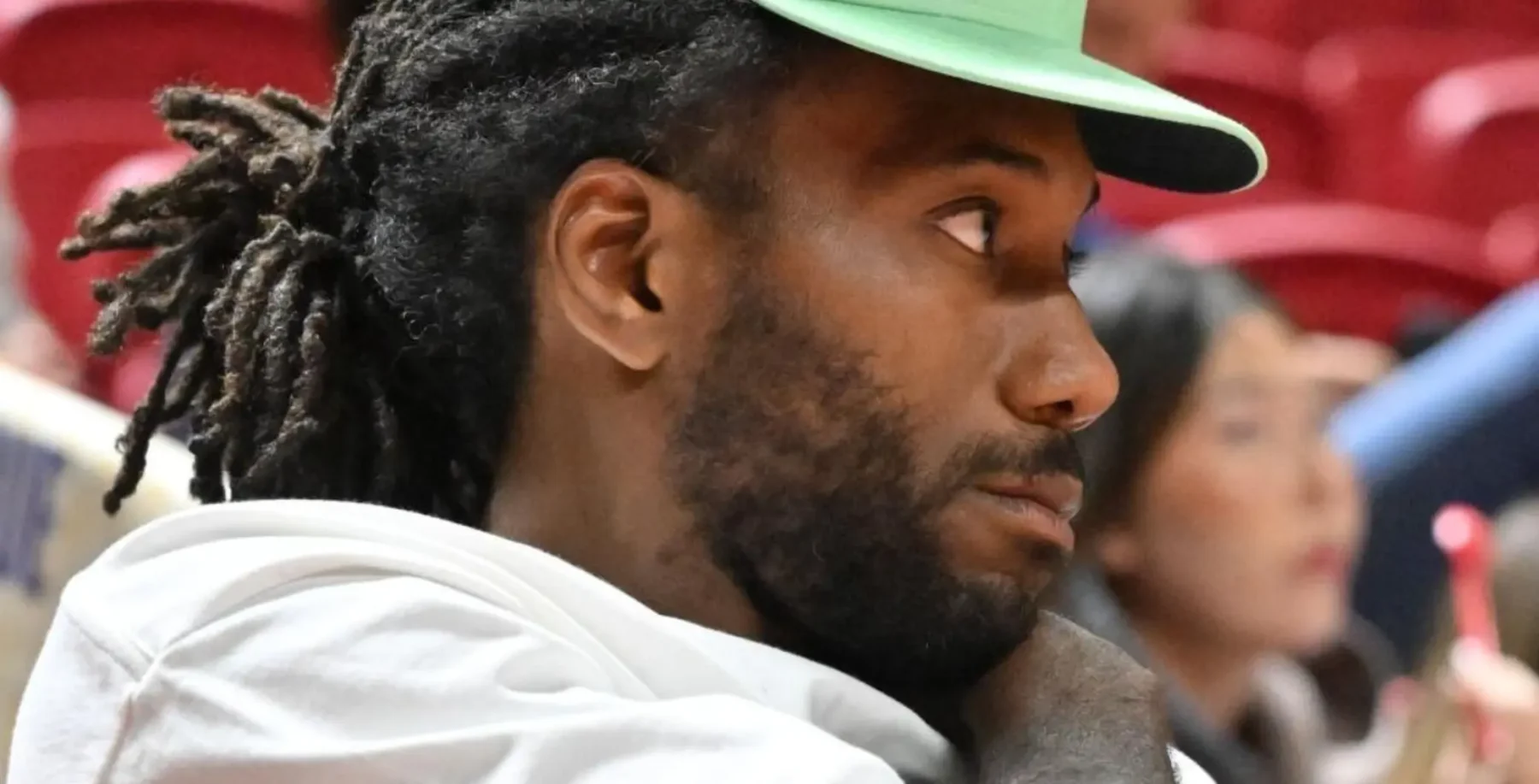 Kawhi Leonard with dreadlocks and a green cap, sitting courtside at a Toronto Raptors game, focused on the action.
