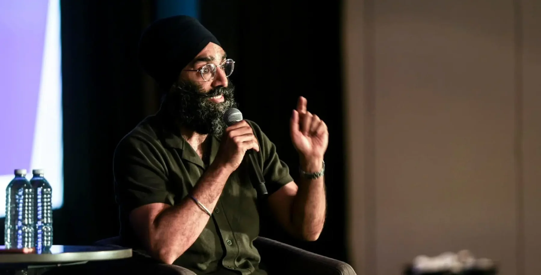 Jagmeet Singh's lawyer brother speaking at an event, wearing a turban and glasses, holding a microphone, with a focused expression.