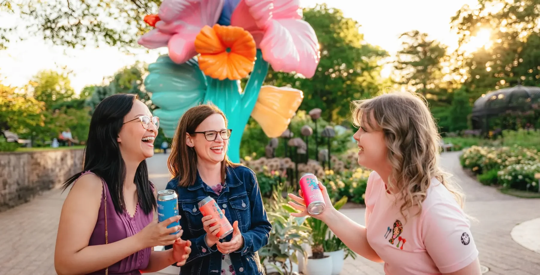 Three women enjoying drinks and laughing outdoors at a park with colorful balloons and lush greenery in the background.