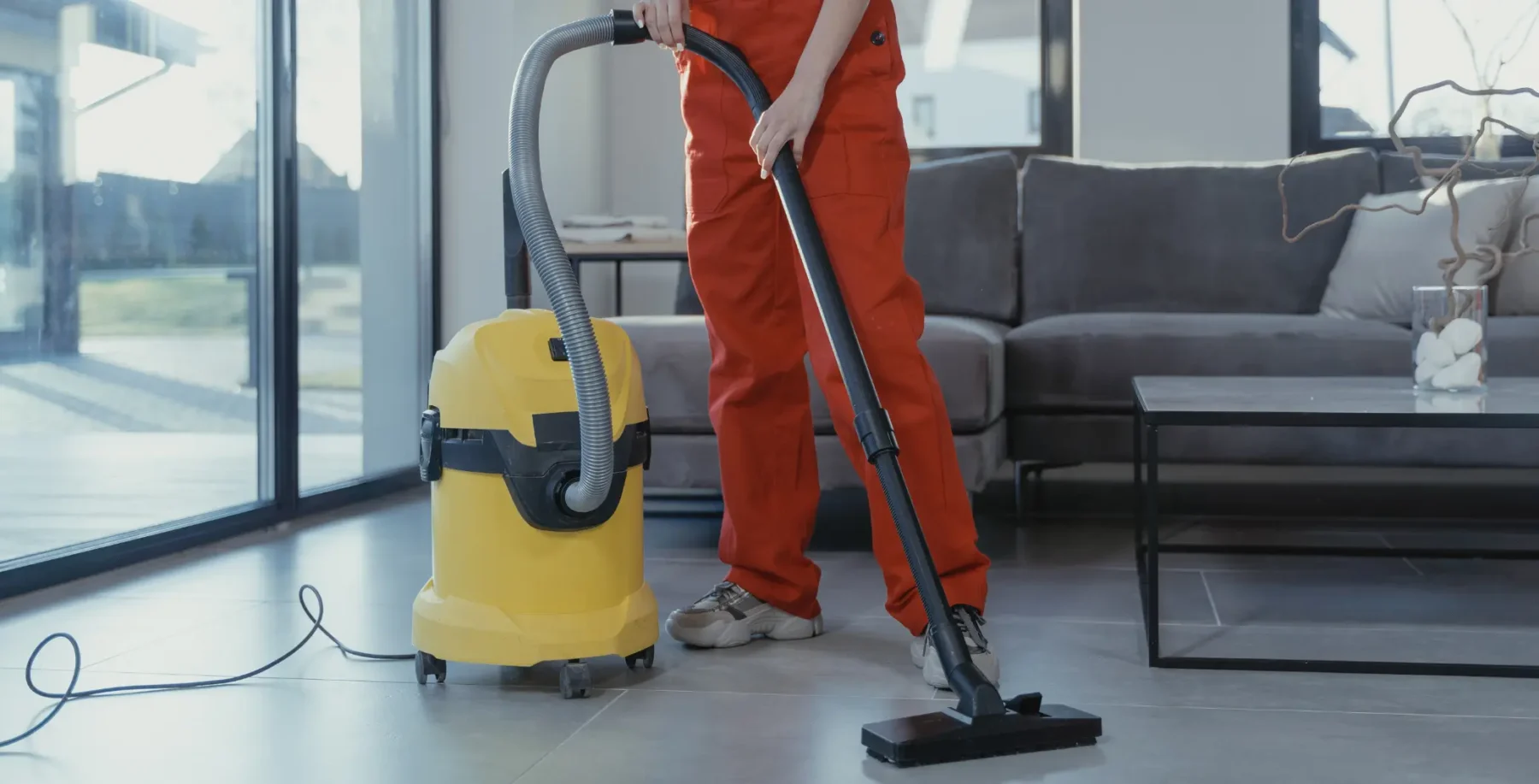 Person using a vacuum cleaner to clean a modern living room with large windows and a gray sofa.