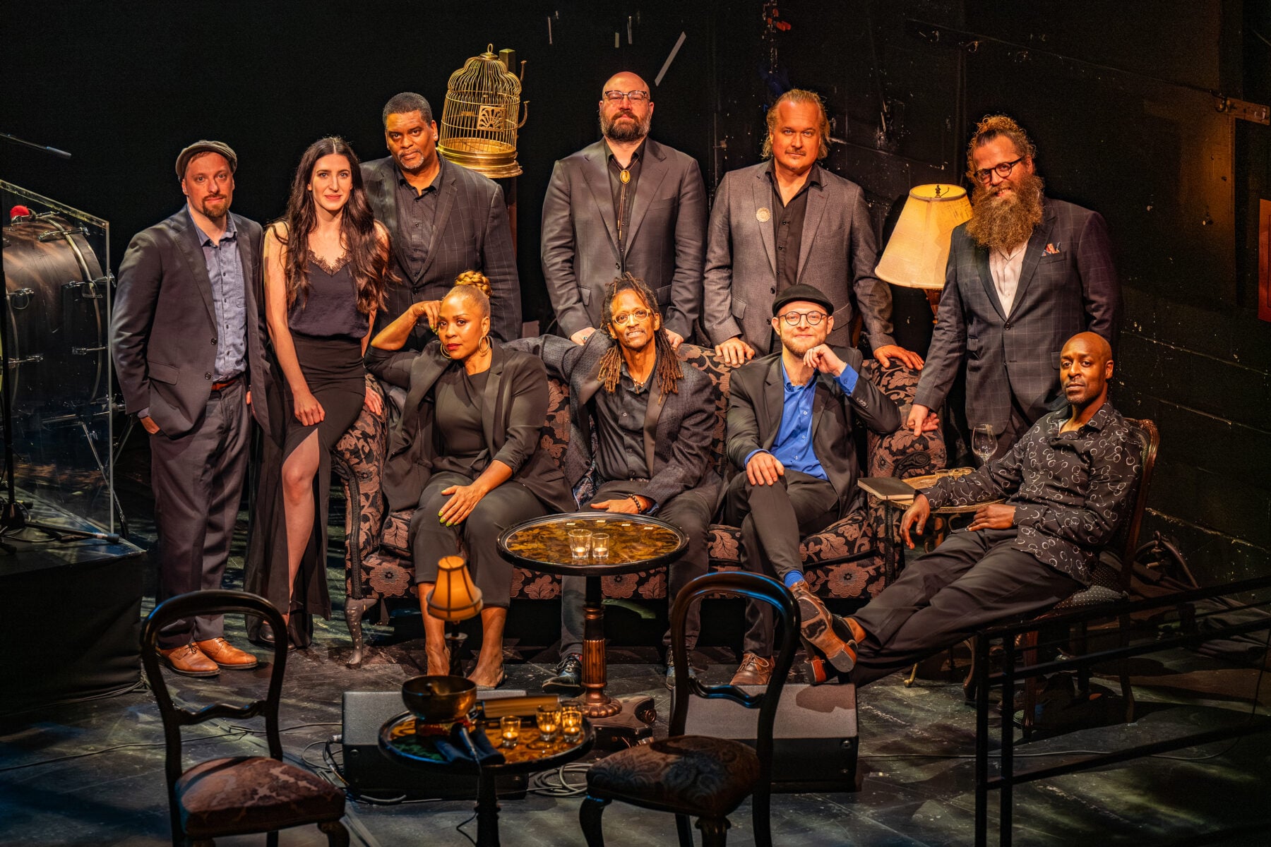 Group photo of cast and crew from Soulpepper's The Secret Chord, a Leonard Cohen experience, on stage with vintage furniture and warm lighting.
