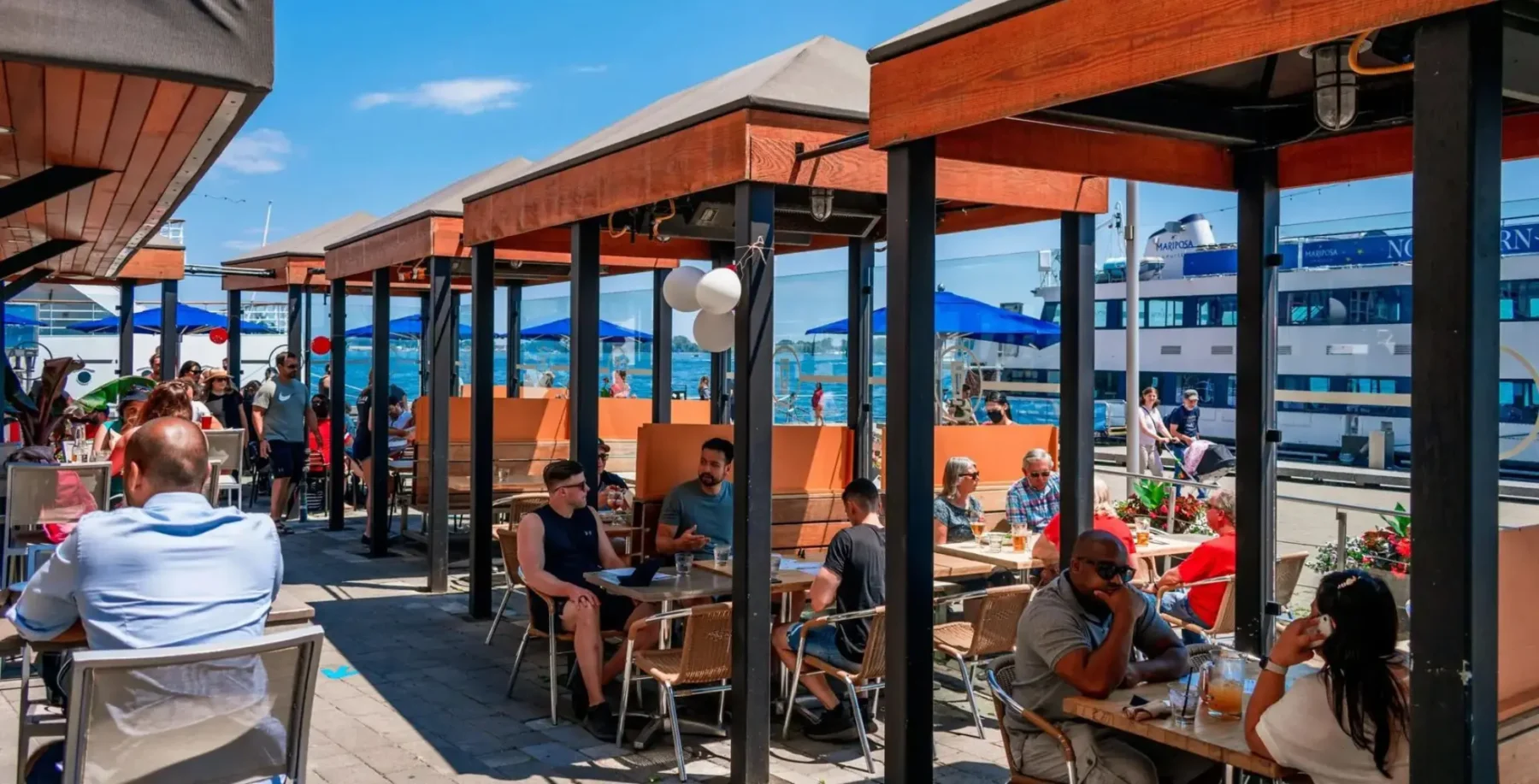People dining outdoors at a Toronto patio with blue umbrellas and a waterfront view, under a clear sky, during summer.