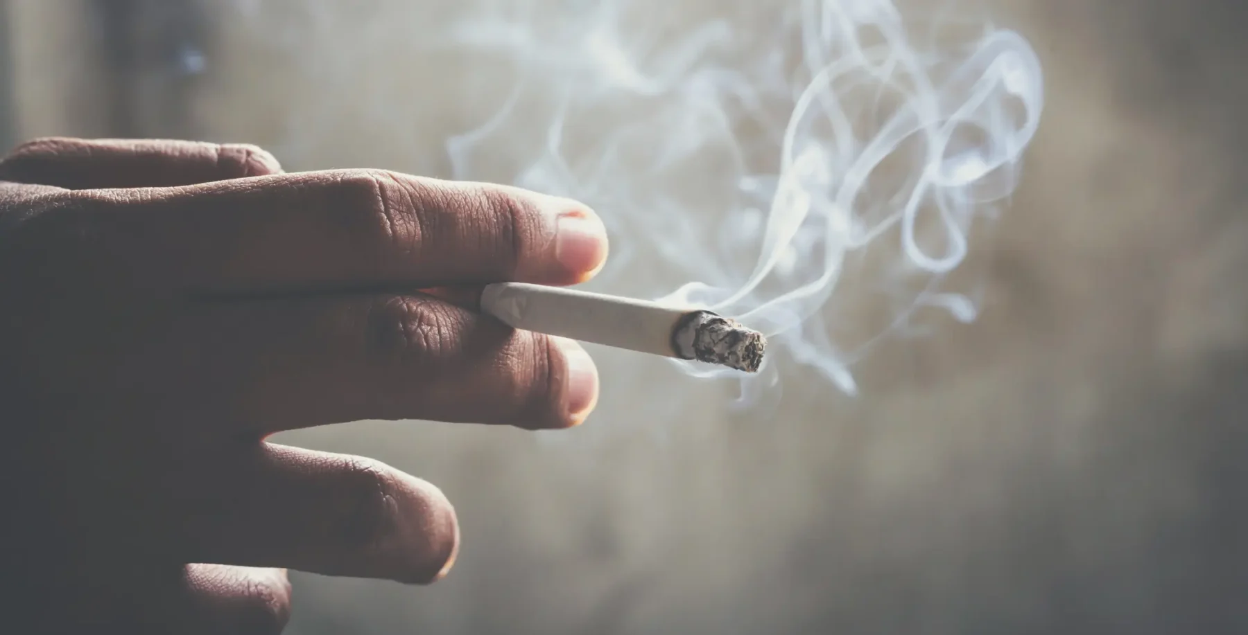 Close-up of a hand holding a lit cigarette with smoke curling up, highlighting health risks of smoking.
