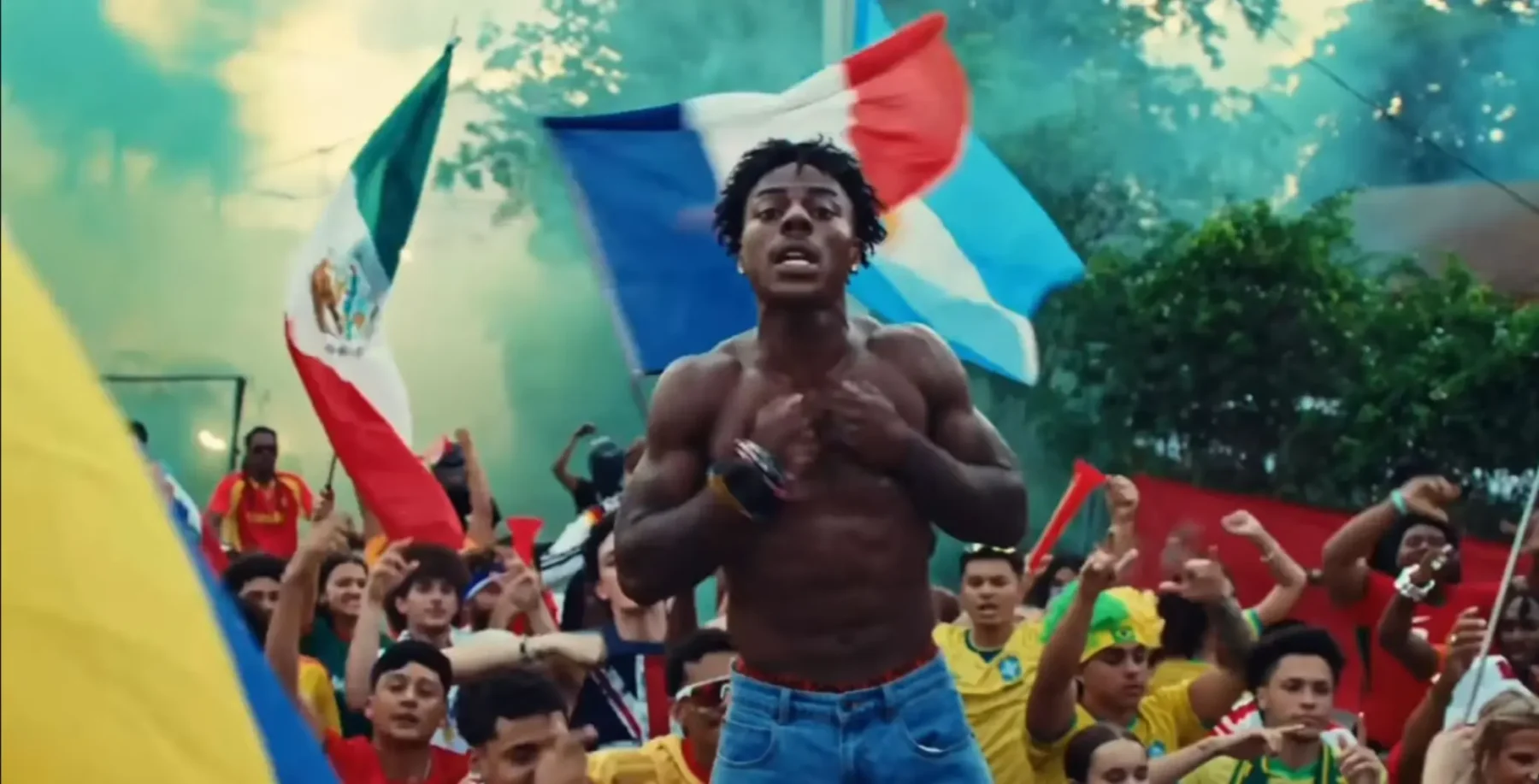 A shirtless young man with curly hair and a wristwatch, holding a flag, surrounded by a crowd of children and adults, celebrating outdoors with flags and smoke.