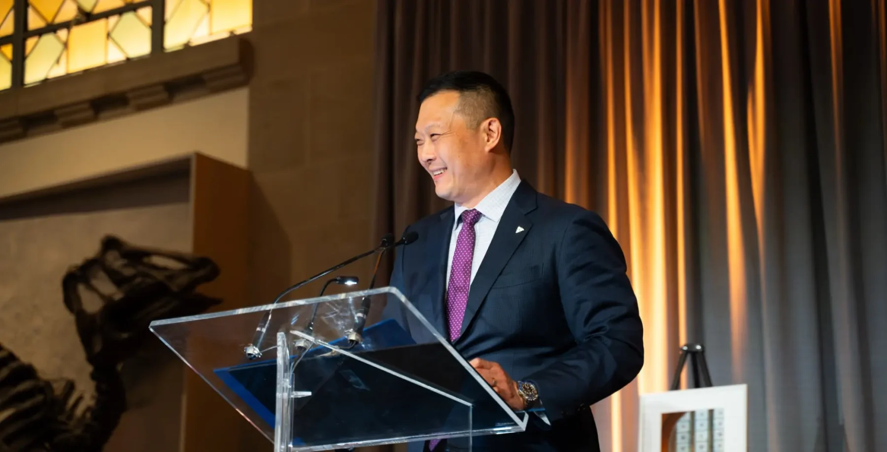 Stan Cho speaking at a podium during a formal event, smiling, with a dark curtain backdrop and warm lighting.