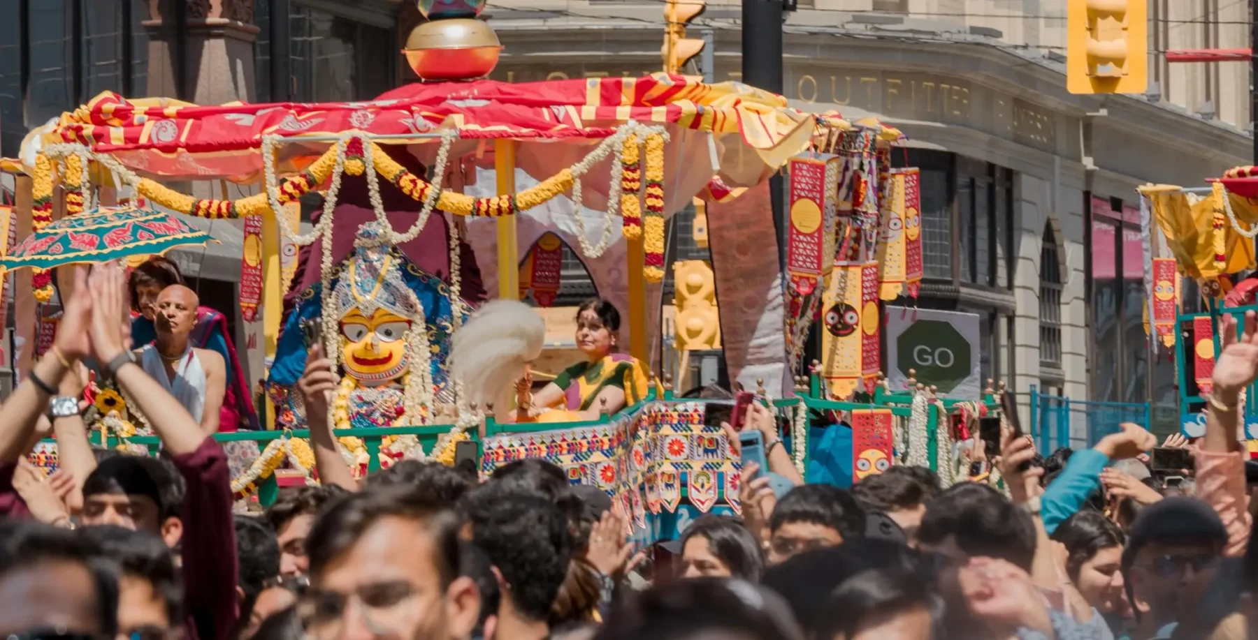 Colorful parade float with traditional decorations and performers during Toronto festival, crowded street scene with spectators and city buildings in background.