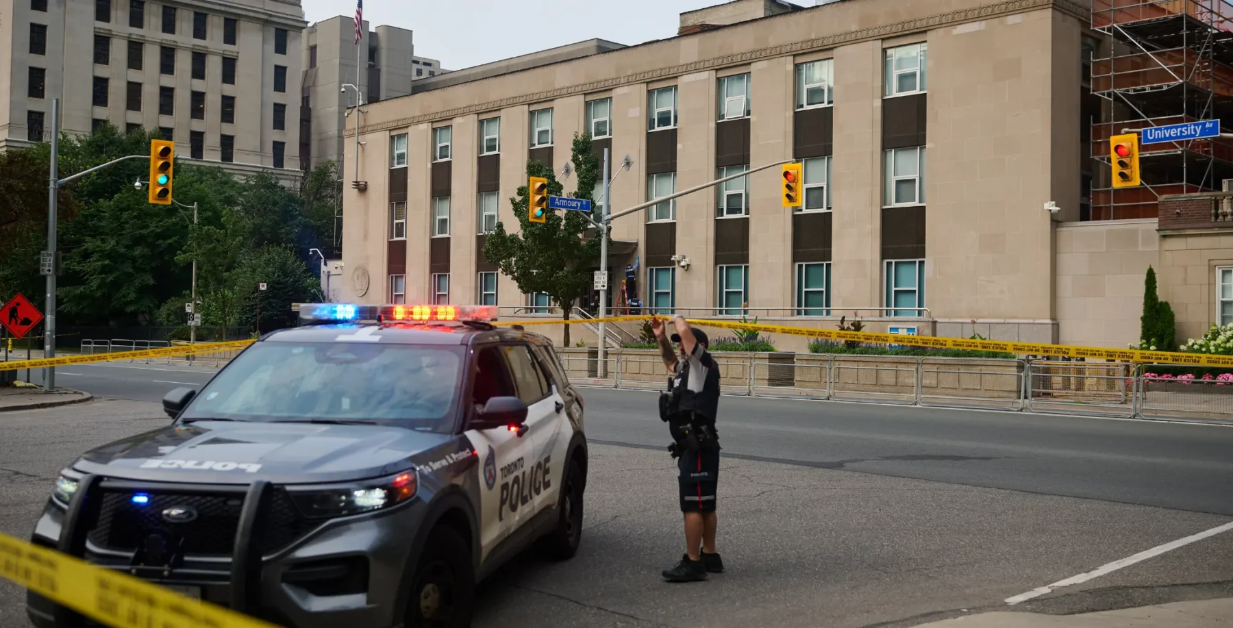 Toronto police investigate a shooting outside the U.S. Consulate, with police car and officer on scene, yellow caution tape, and city buildings in the background.