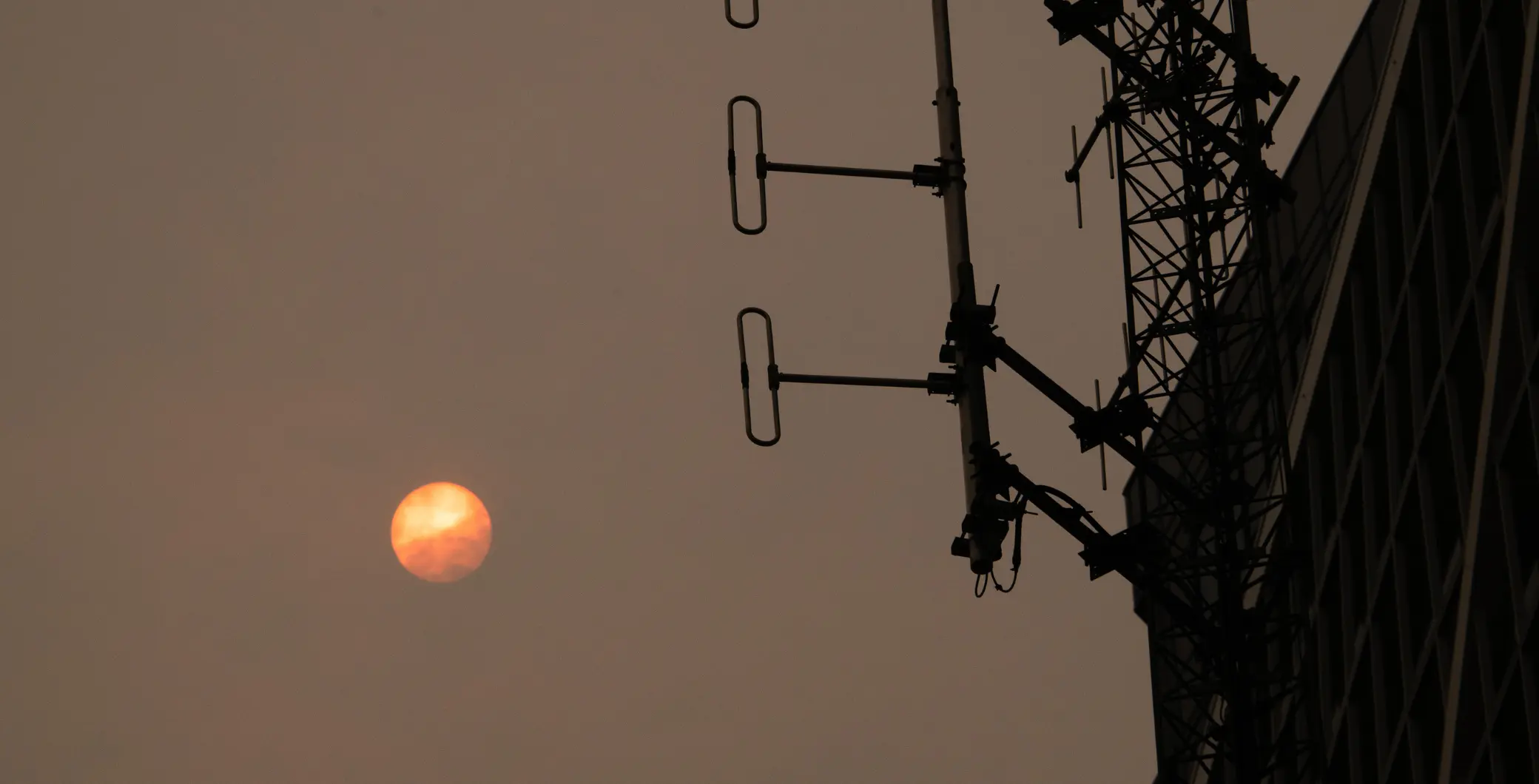 A large, reddish moon visible in the sky near a tall electrical tower with antennas, against a dusky, orange-tinted sky in Toronto.