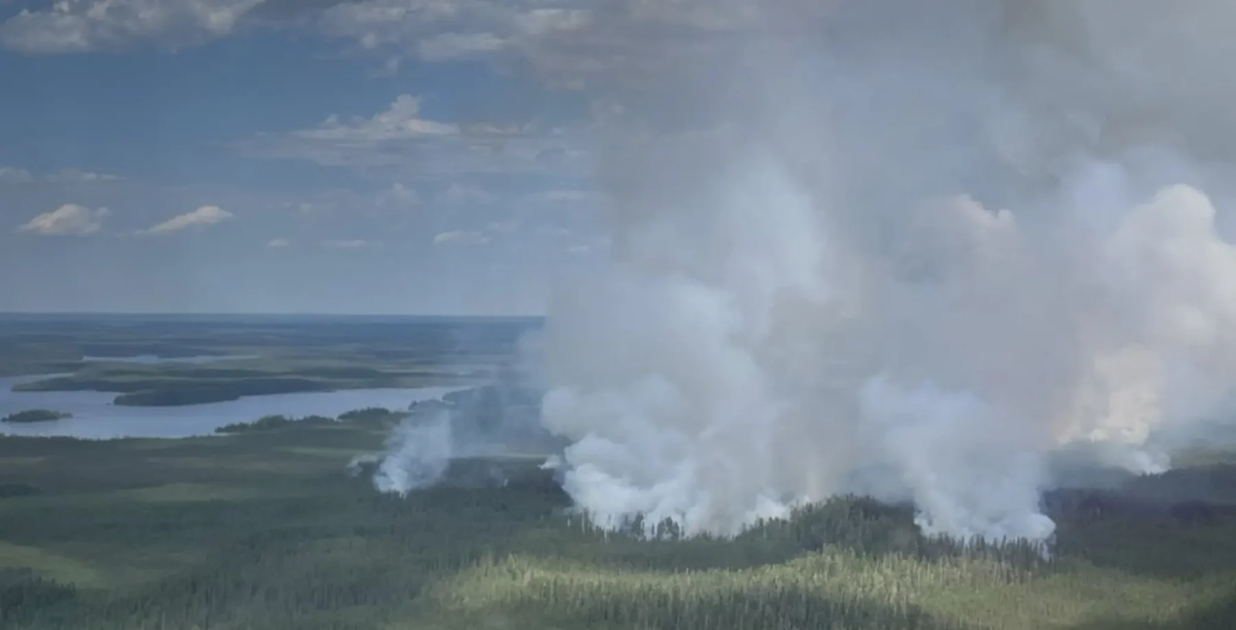 Smoke rising from a wildfire in a forested area near a body of water, with a cloudy sky overhead.