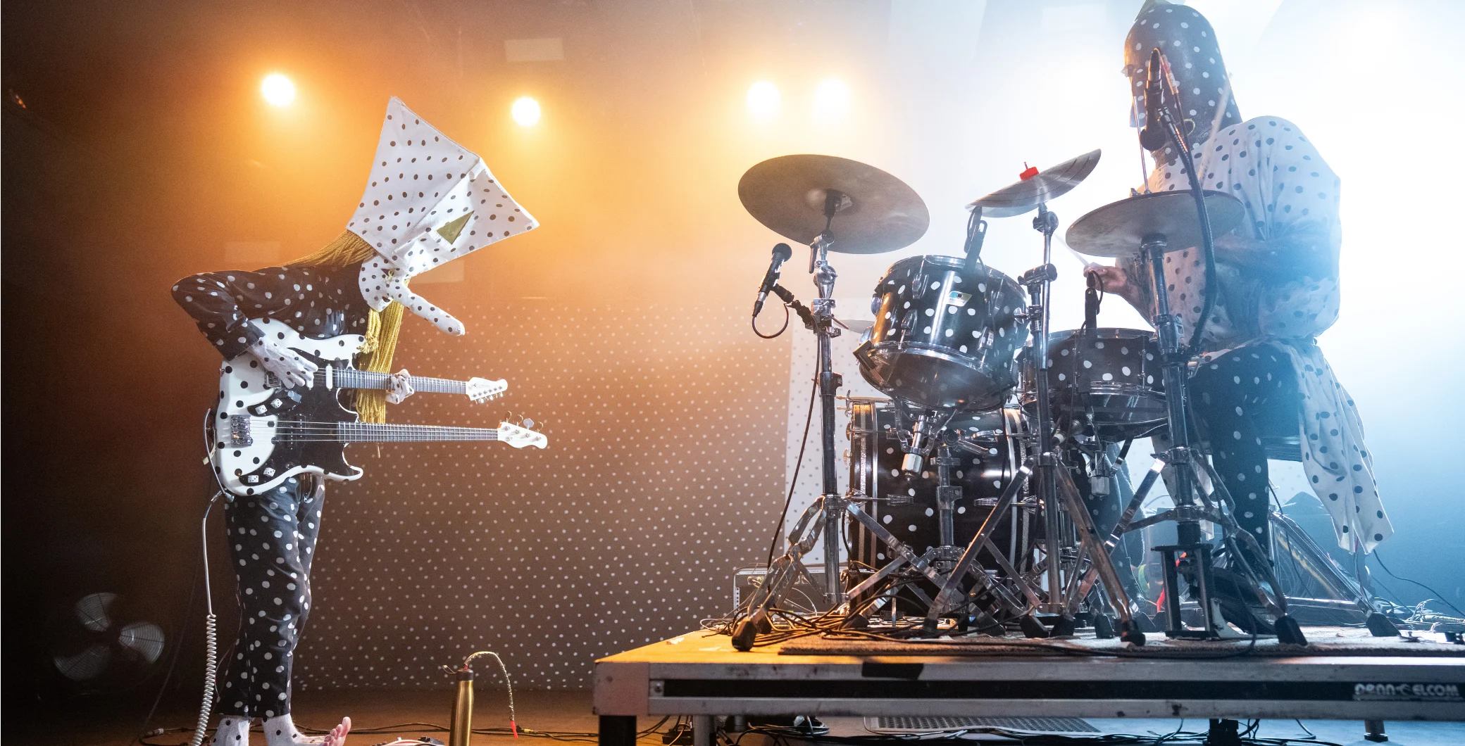 Two musicians dressed in polka dot costumes perform on stage with guitars and drums, illuminated by warm stage lighting at a live concert.