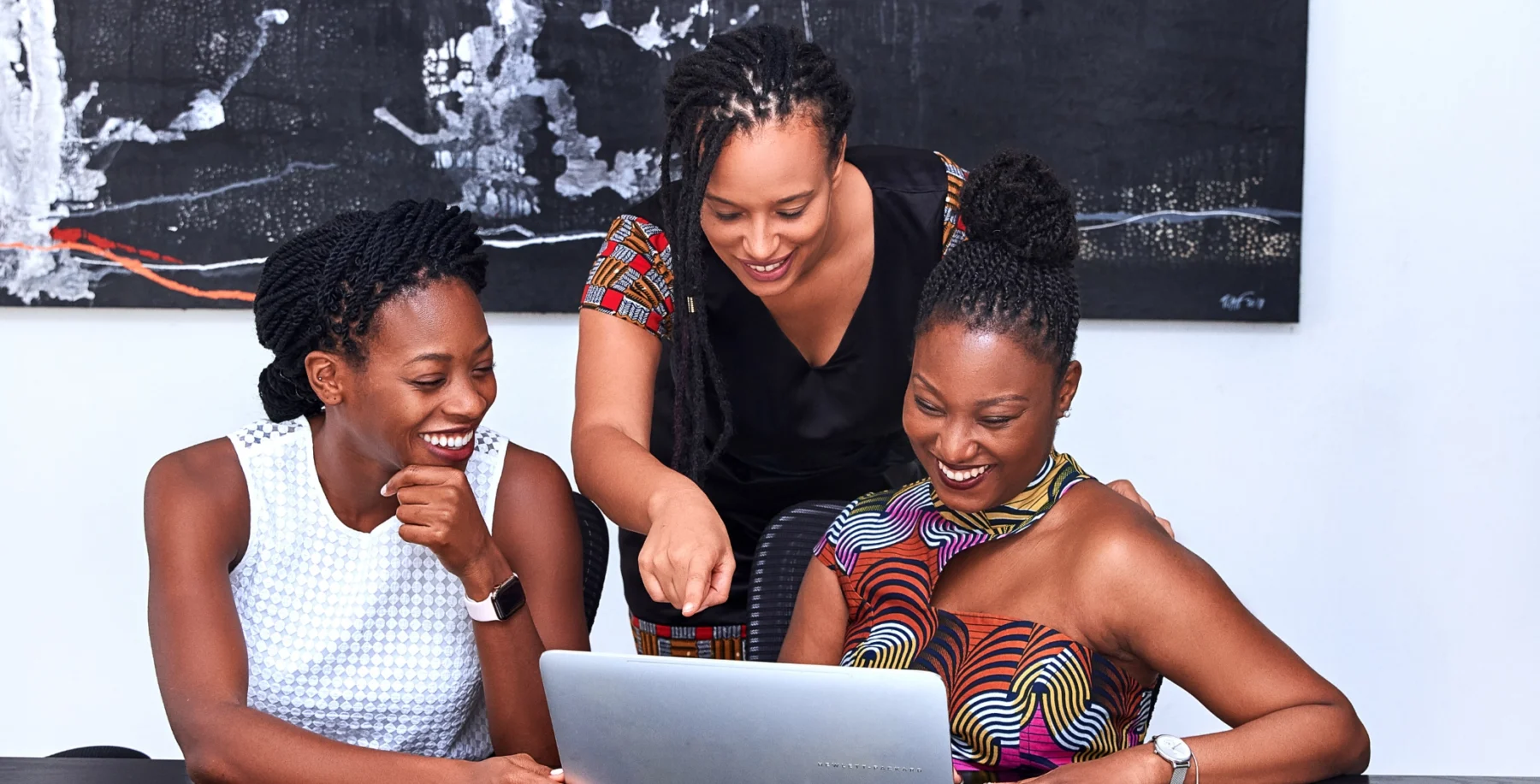 Three women of diverse backgrounds smiling and collaborating around a laptop in a modern office setting.