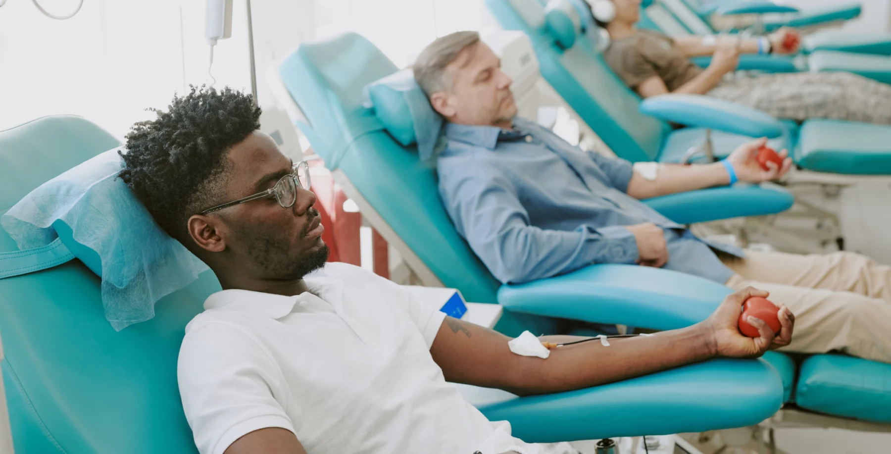 People donating blood at a medical clinic, lying on chairs with blood collection tubes and red stress balls in hand.