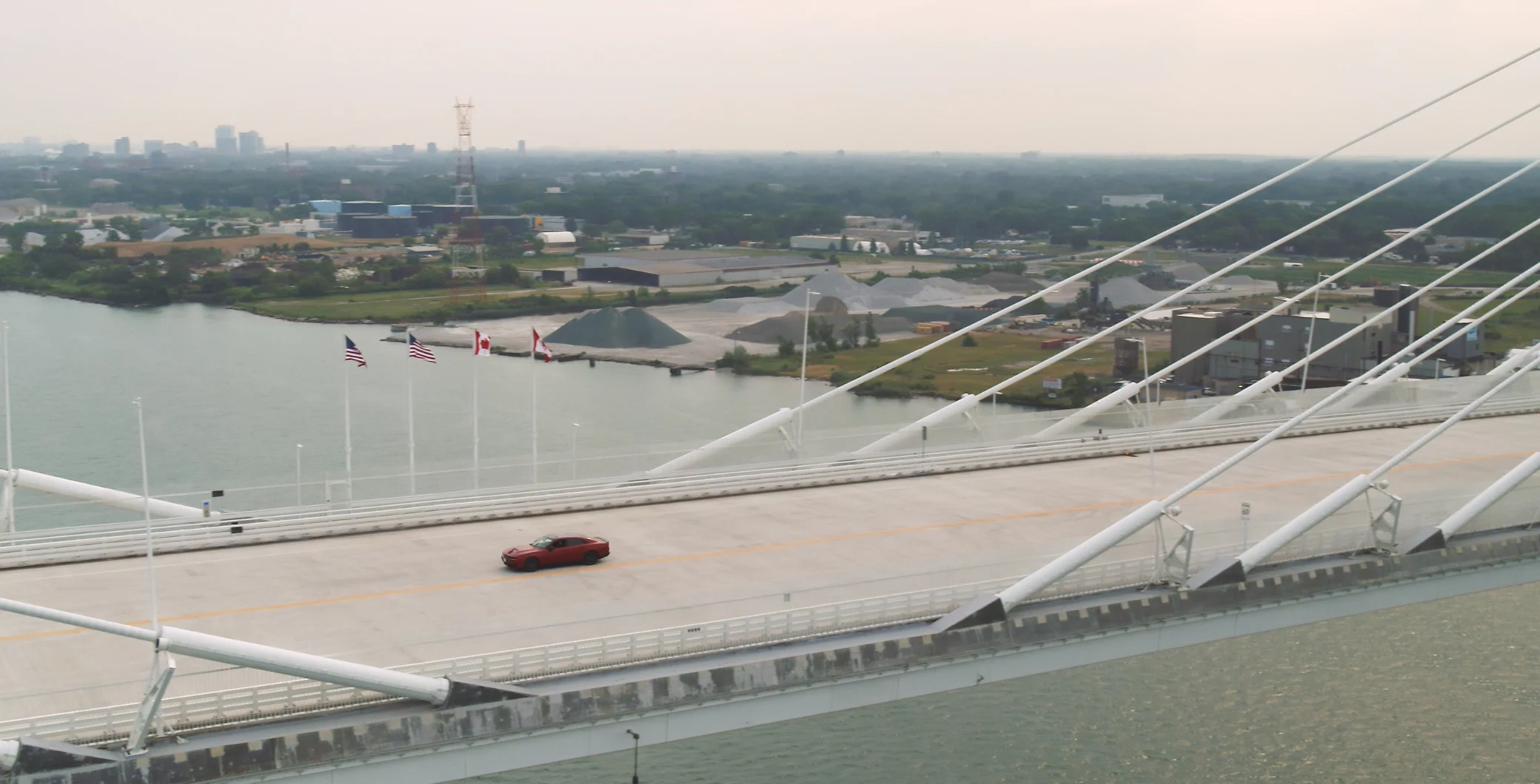 A red car driving across a modern cable-stayed bridge over a river in Toronto, with cityscape and industrial areas in the background.