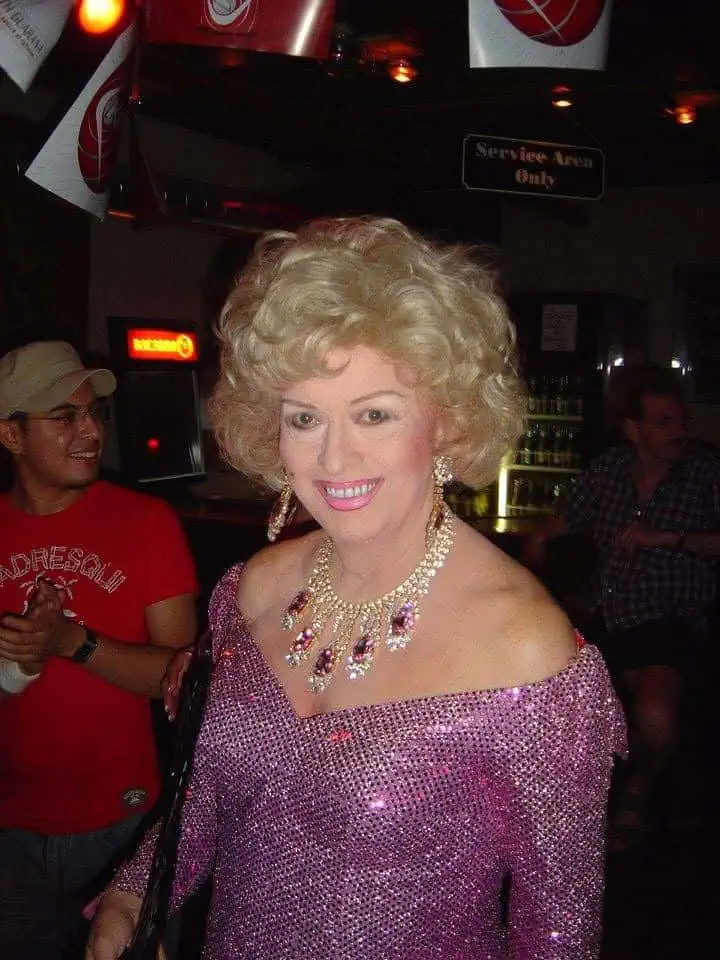 A smiling woman with curly blonde hair wearing a pink sequined dress and jewelry at a lively indoor event with people in the background.