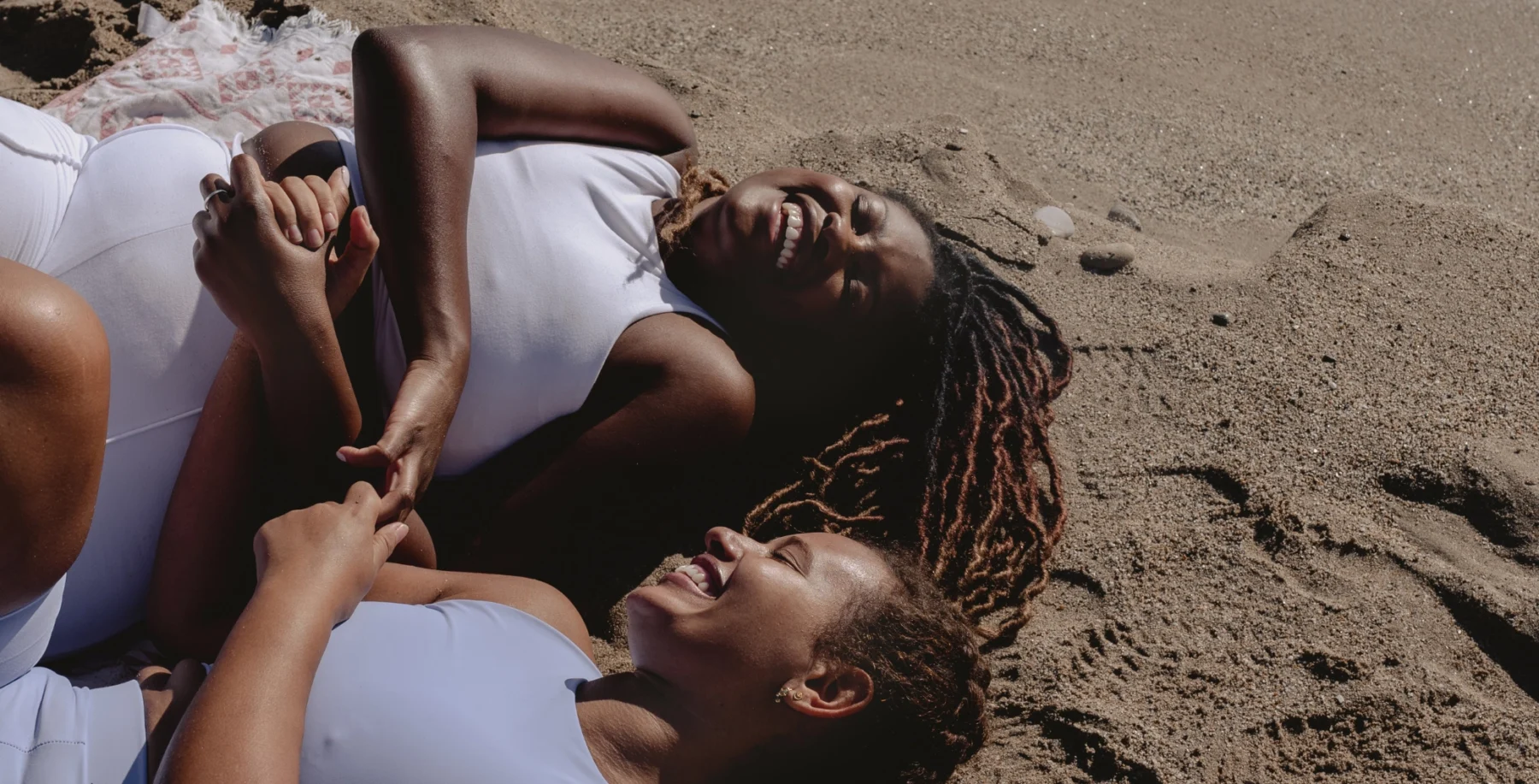 Two BIPOC women relaxing and laughing on a sandy beach, celebrating inclusivity and queer culture at Toronto's historic Queer beach.