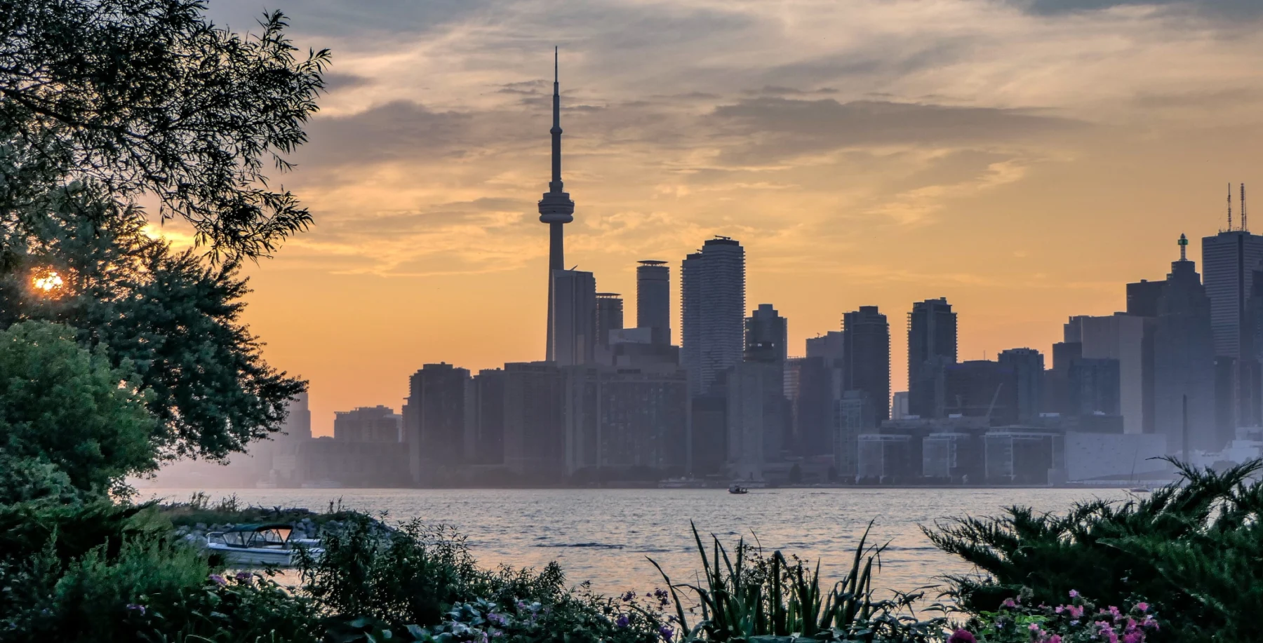 Toronto skyline at sunset with CN Tower and city buildings visible from a park with trees and water in the foreground.