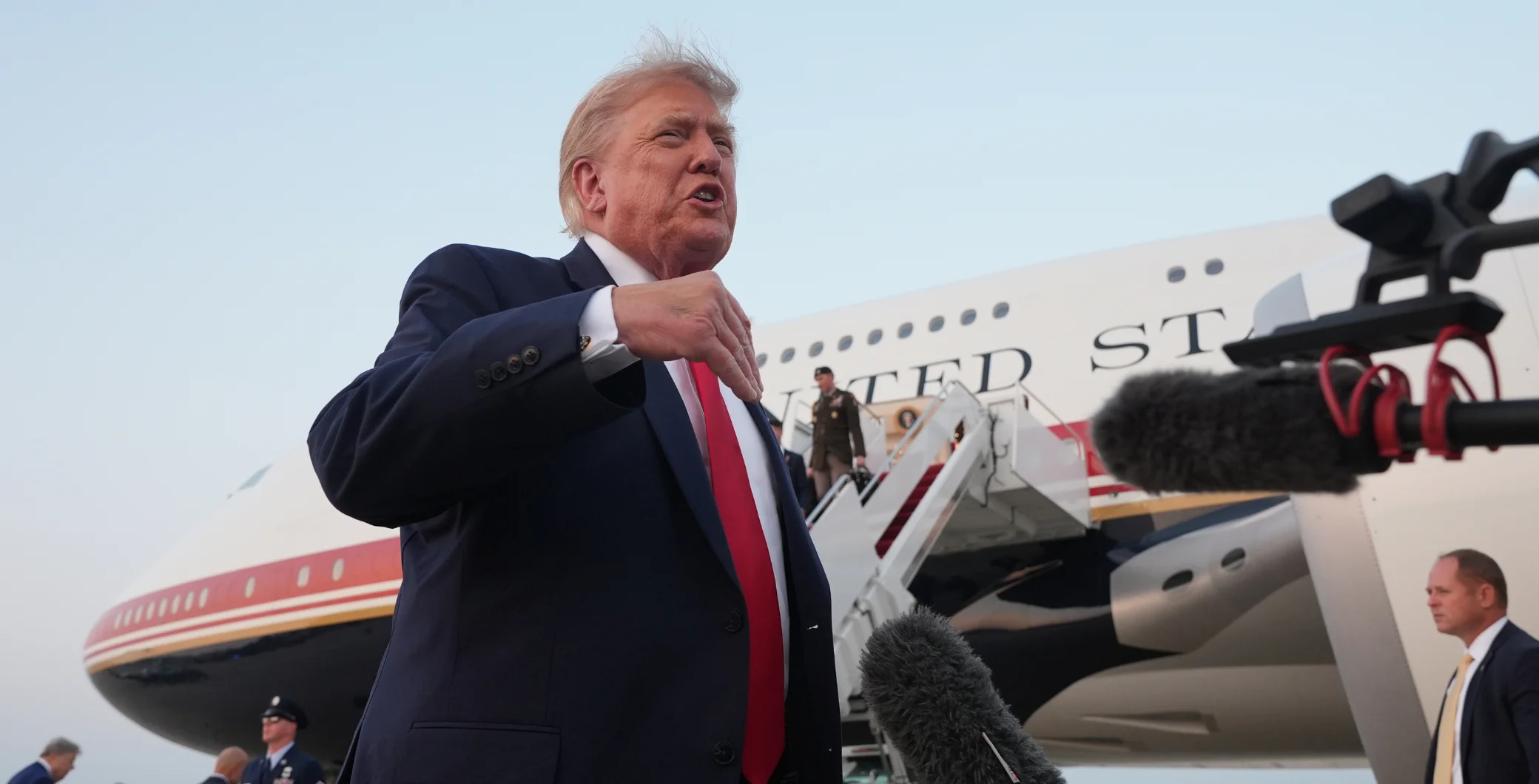 Donald Trump speaking at an airport tarmac with a private jet in the background, surrounded by media microphones and security personnel.