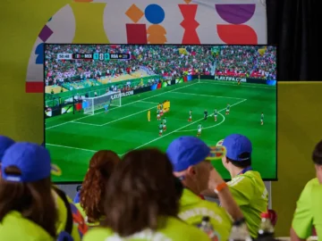 Children watching FIFA World Cup match on a large screen at a watch party in Toronto, wearing blue caps and bright yellow shirts.